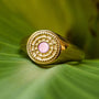 "Pixa Ring", a gold ring featuring pink nacre with intricate circular design on a green leaf backdrop.