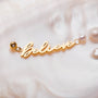 "Creer-Cámara" encanto oro con perlas, con un diseño cursivo "Creer" en un fondo suave.