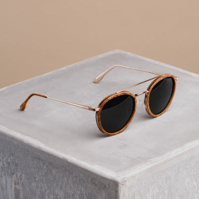 Les lunettes de soleil "The Enchanting" avec un cadre en bois de cannelle et des accents en or mat sur un fond en béton minimaliste.