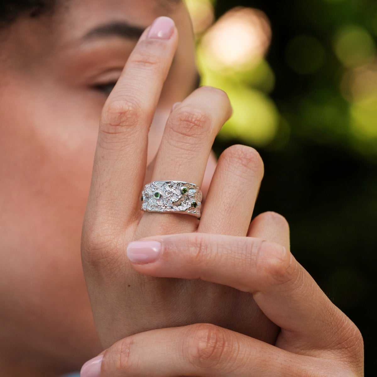 "Silverstream Ring", a silver ring with a textured design and green diopside accents, showcased on a hand against a blurred background.