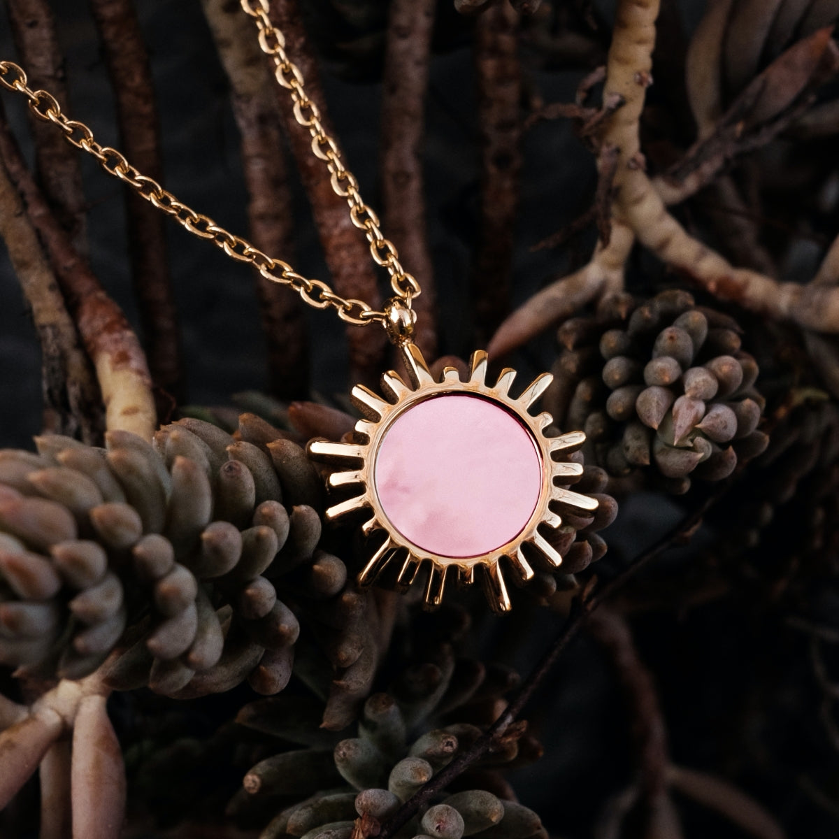 "Shade Necklace", a necklace featuring pink nacre and gold, with a radiant, sun-like pendant nestled among textured flora.