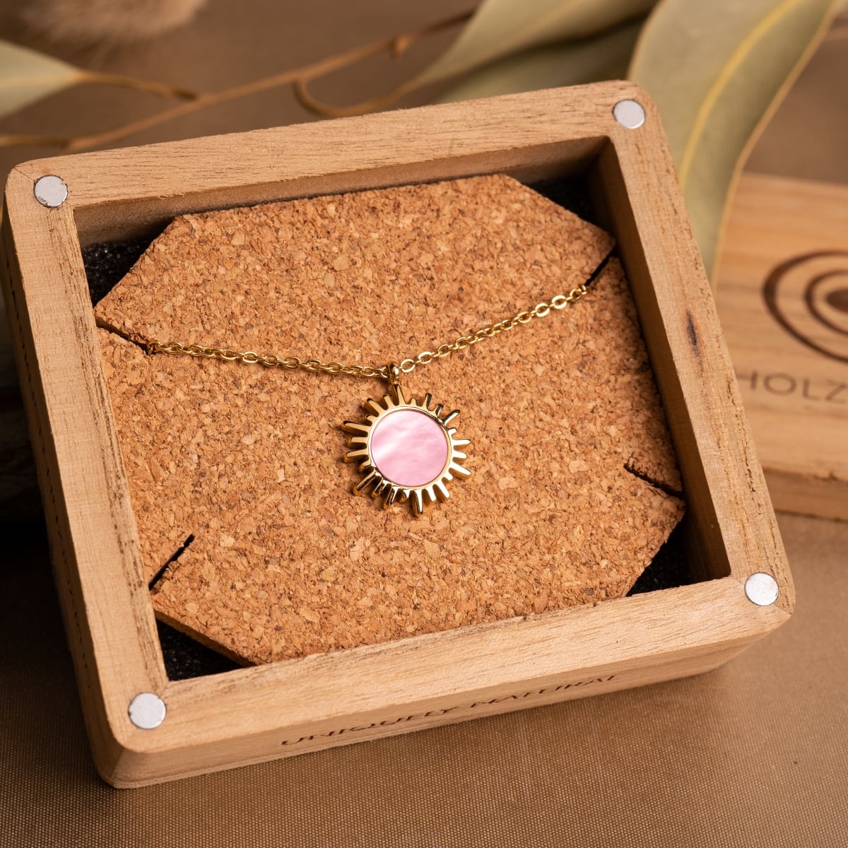 "Shade Necklace" necklace, featuring a sun-themed pink nacre pendant and gold chain, elegantly displayed in a wooden box.