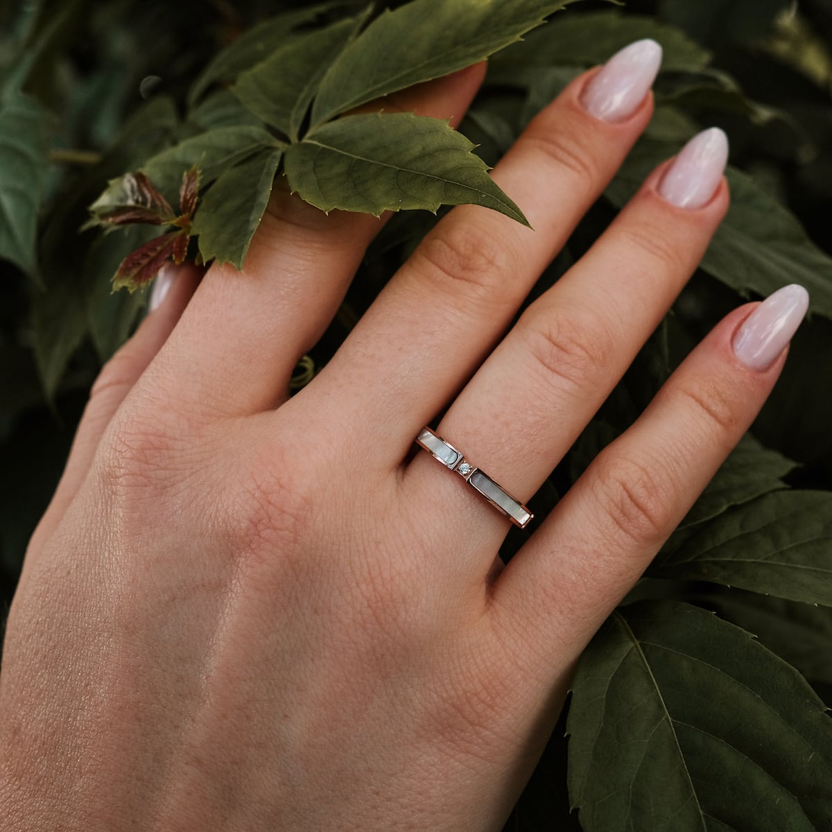 "Campania Ring", une bague élégante avec des accents nacre et or rose, montrée sur une main au milieu de feuilles vertes luxuriantes.